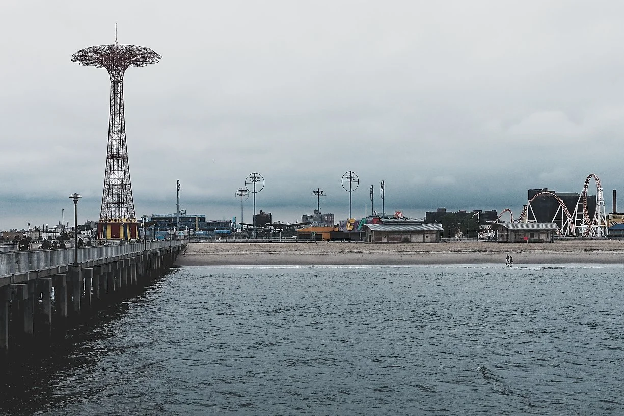 A pier extending into water with a cityscape in the background, featuring a large tower, amusement park rides, and several streetlights, on an overcast day.