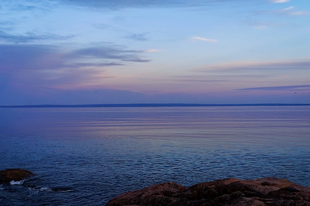 Et rolig hav med lave bølger under en blålig himmel med spredte skyer, sett fra en kyst med større steiner i forgrunnen.