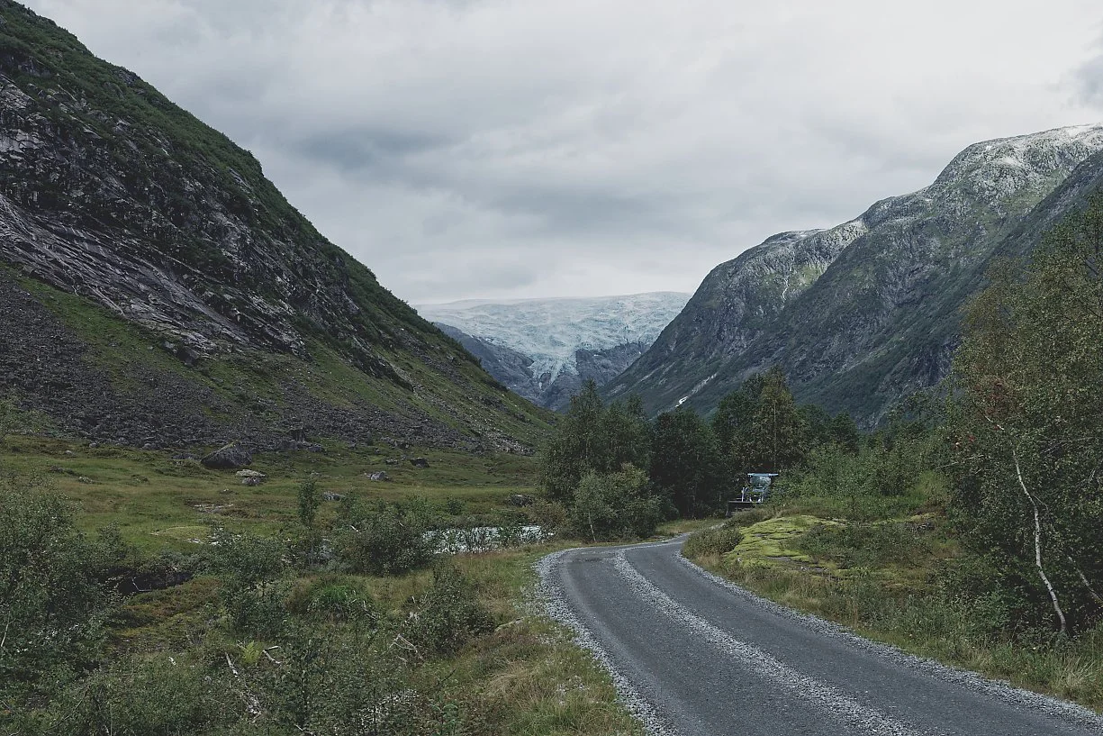 En grusvei som slynger seg gjennom en fjellrik dal med grønne busker og trær, omgitt av høye, steinete fjell med snø på toppene, under en overskyet himmel.