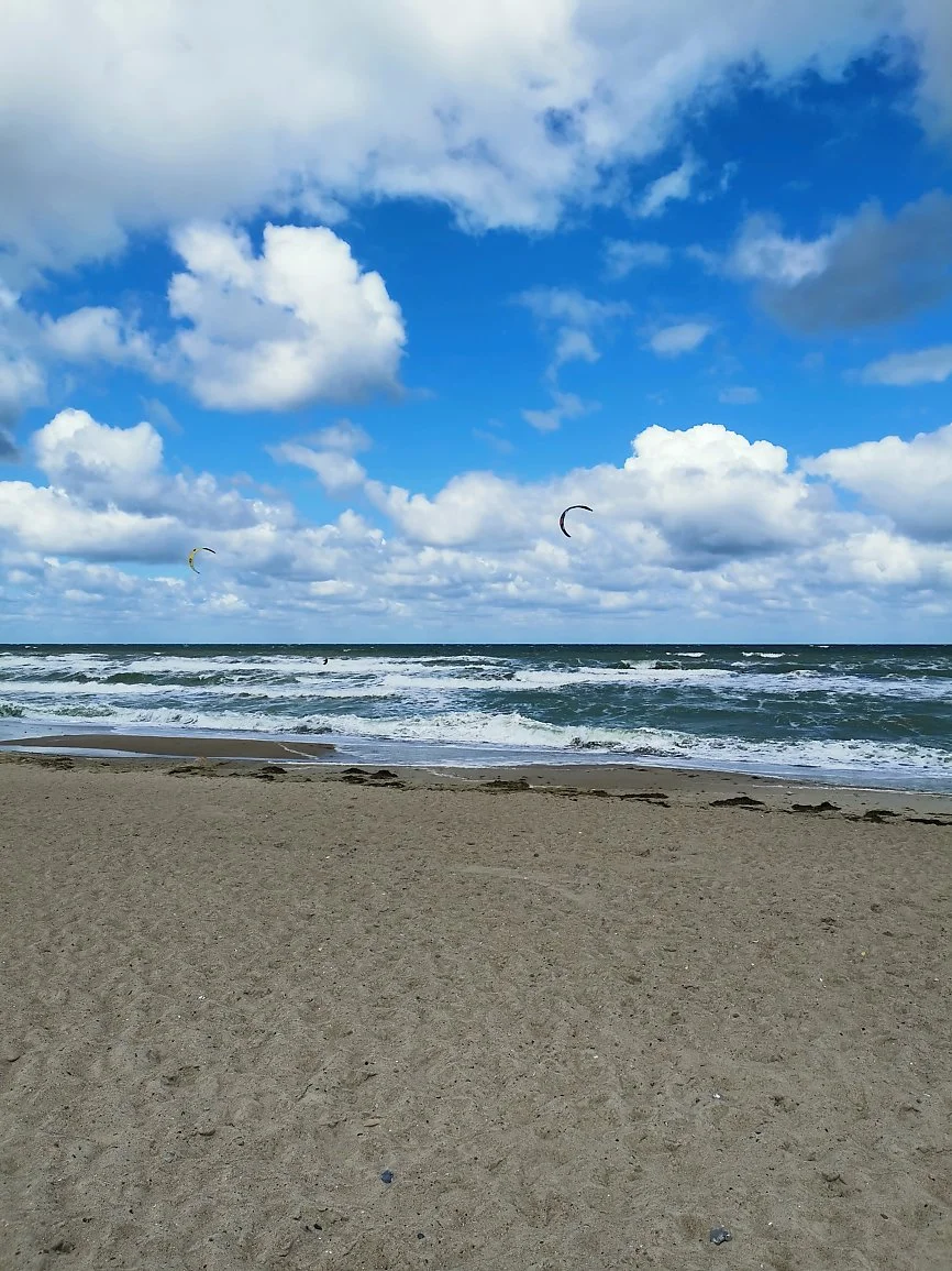 A beach scene with sandy shore, ocean waves, and a blue sky with clouds. Two kite surfers are visible with their kites in the sky.
