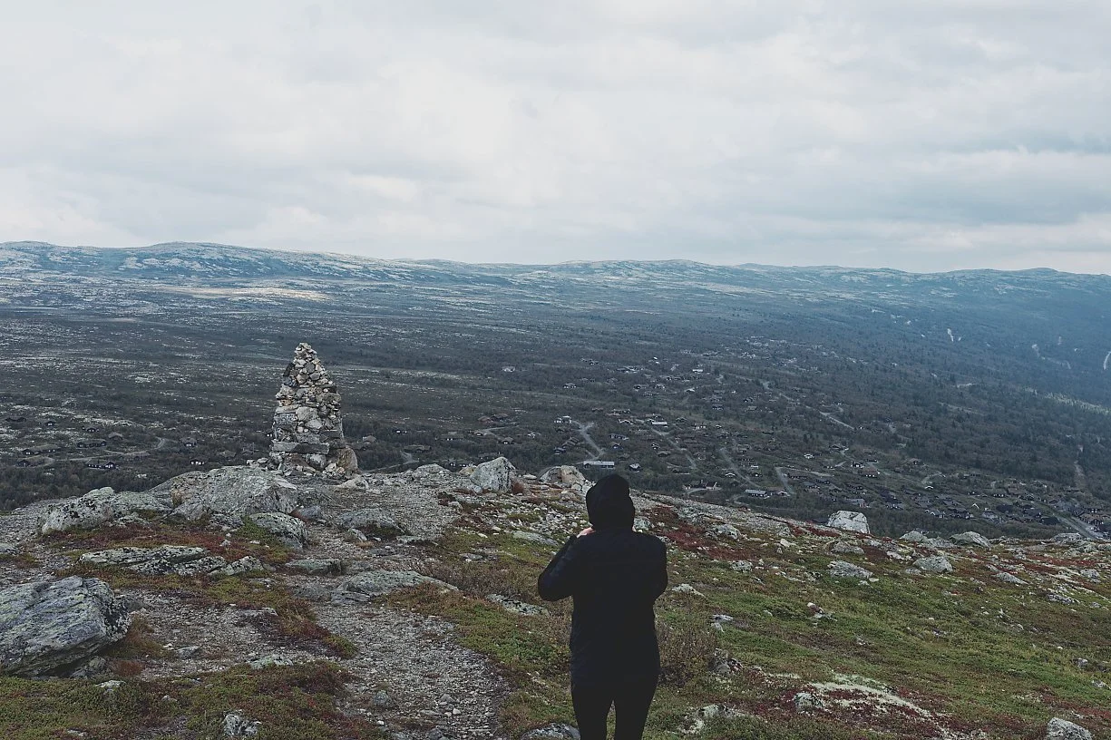 Person som står på en fjelltopp med utsikt over et landskap med åser og en liten landsby, under en overskyet himmel.