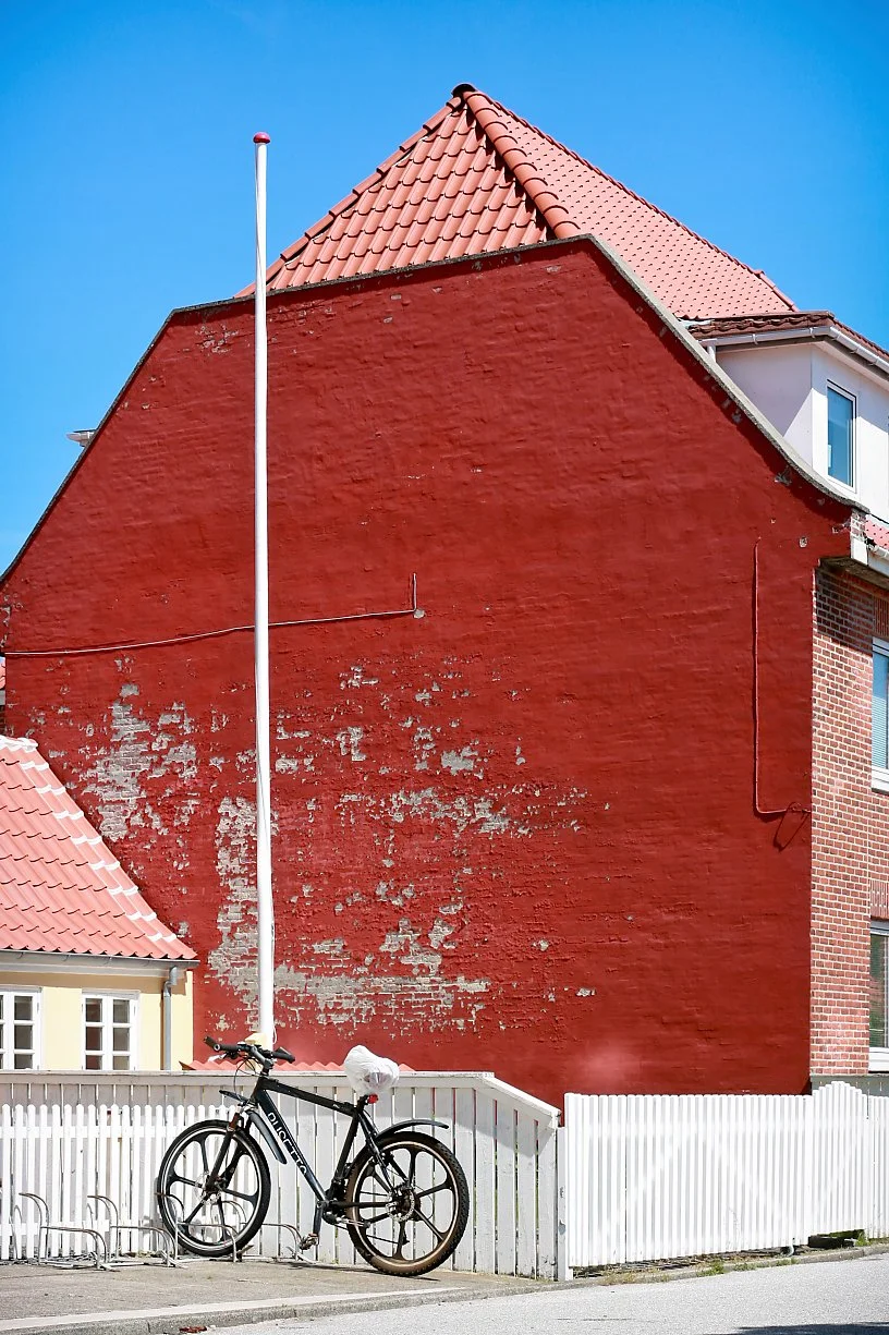 A black bicycle leaning against a white fence near a large red brick building with peeling paint, and a light red tiled roof, under a clear blue sky.