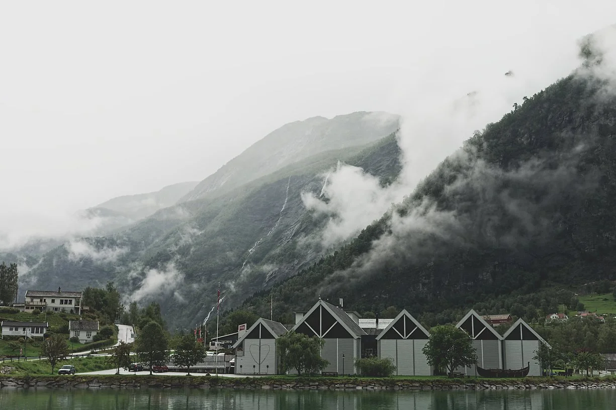 En dal med fjell og skog, med lave skyer og tåke som ligger over fjellene. I forgrunnen er det en liten by med hus og en sjø eller en innsjø.