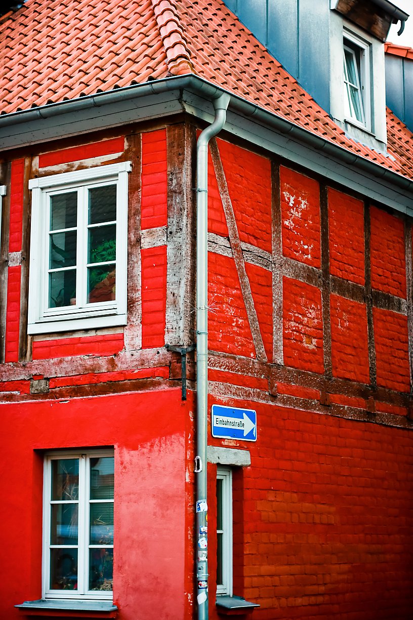 Close-up of a red brick building with white-framed windows, wooden beams, and a red tiled roof. A street sign in German is attached to a pole in front of the building.