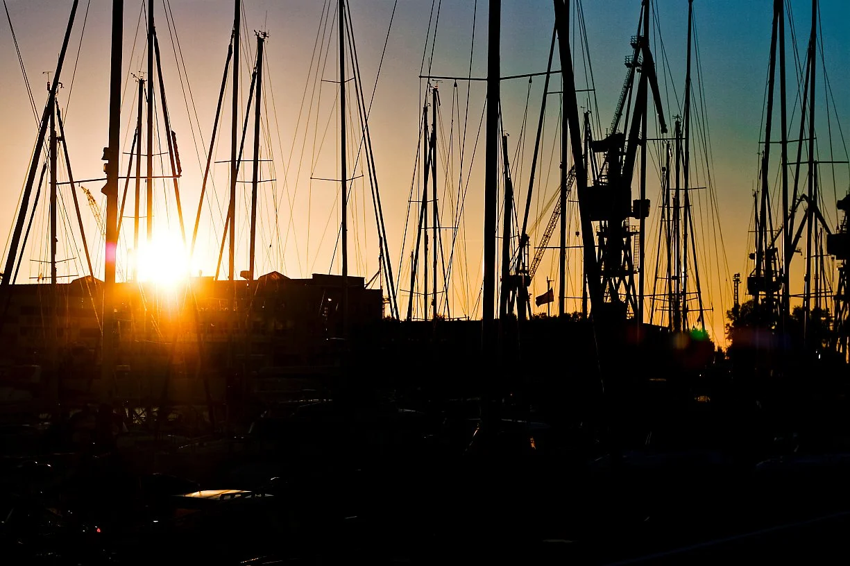 Silhouetted sailboats with masts against a sunset or sunrise sky at a marina.