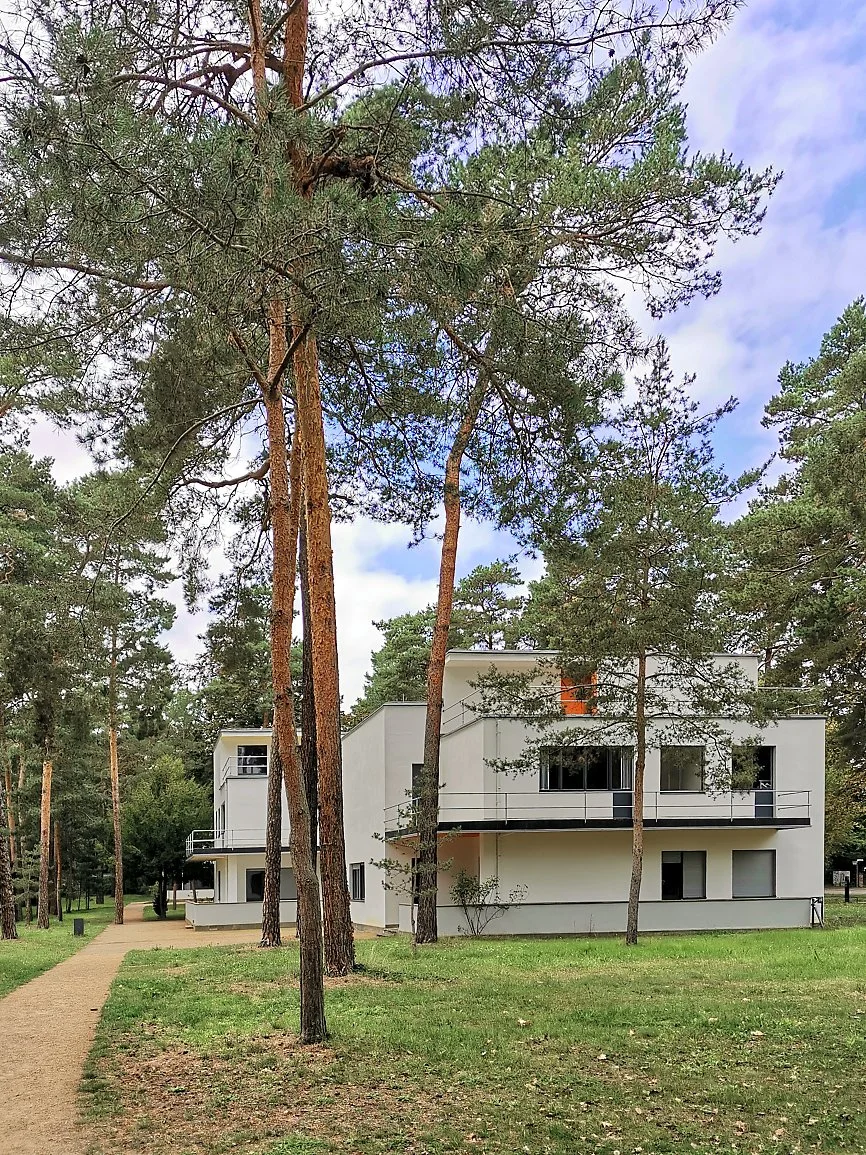 White modern multi-story apartment building with balconies, surrounded by tall pine trees and a grassy park area, under a partly cloudy sky.