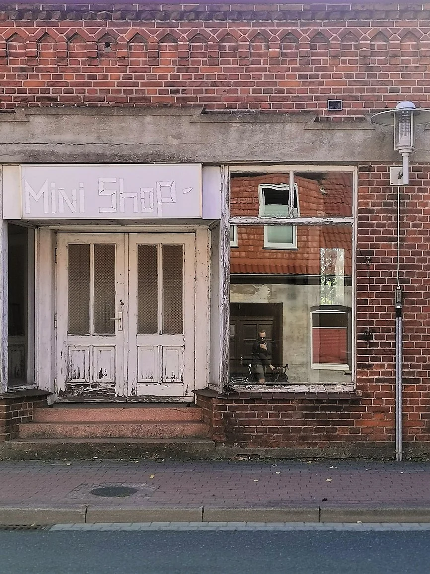 An old brick building with a worn-out white double door and a glass window with reflections, including a person wearing a mask taking the photograph.