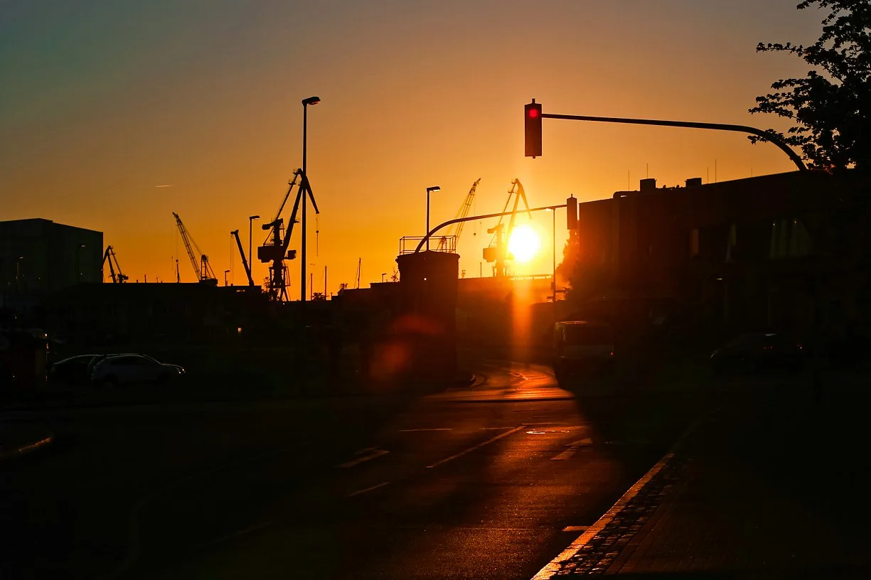 Sunset over a harbor with construction cranes and a traffic light showing red.