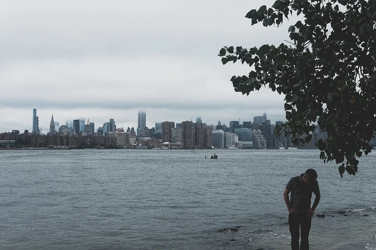 A person standing near the water's edge under a tree, with a city skyline and overcast sky in the background.