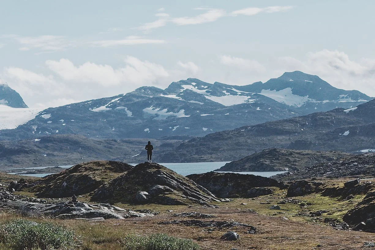 En person står på en klippe i en fjellandskap med snødekte fjell i bakgrunnen og en innsjø i forgrunnen.