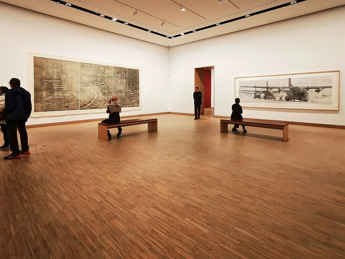 Innvendig på kunstmuseum med fire personer som ser på kunstverk på veggene, noen sitter på benker, og det er tre store bilder på veggene.