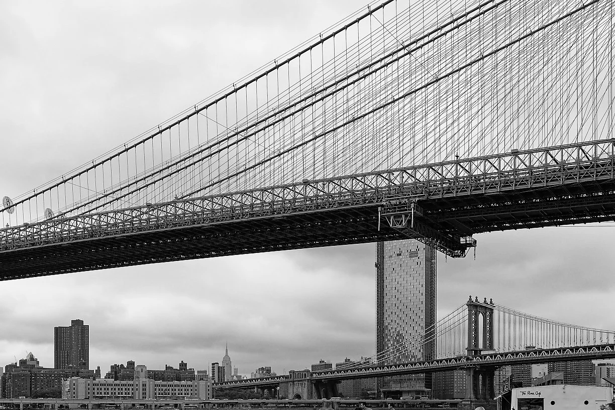 Black and white photo of a bridge with a city skyline in the background, featuring tall buildings including the Empire State Building.