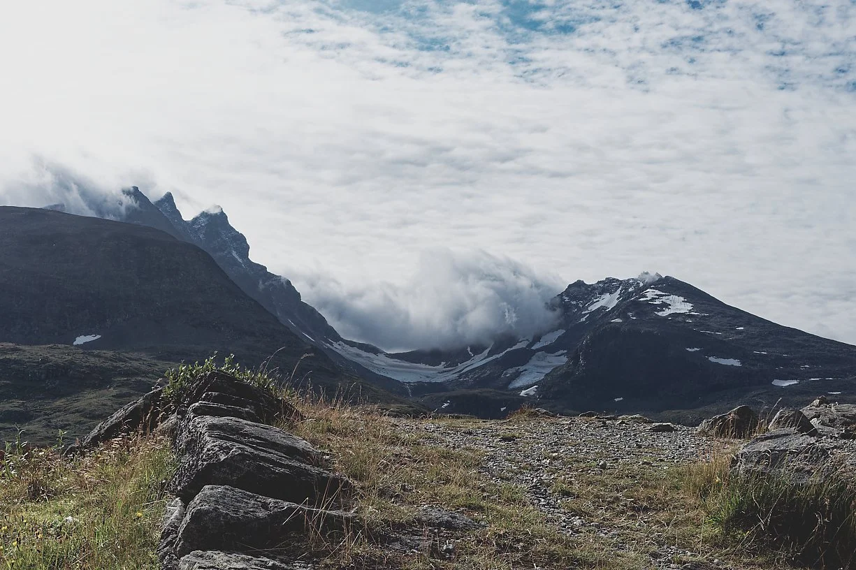 Fjellandskap med snødekte fjell, skyer og en sti med steiner i forgrunnen.