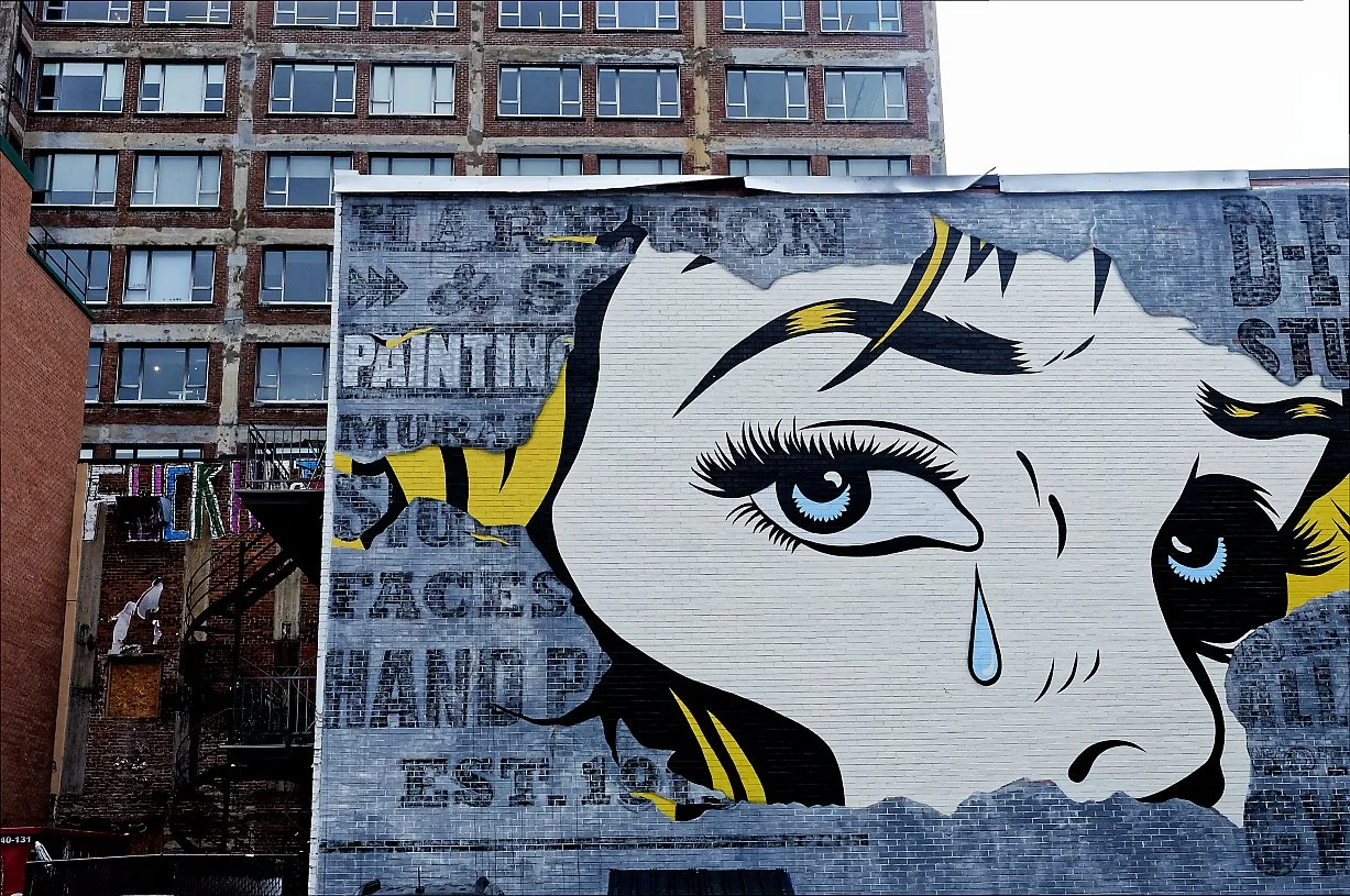 Street artwork featuring a large portrait of a girl with blue eyes and a teardrop, painted on a building wall with partially visible graffiti and surroundings of urban buildings.