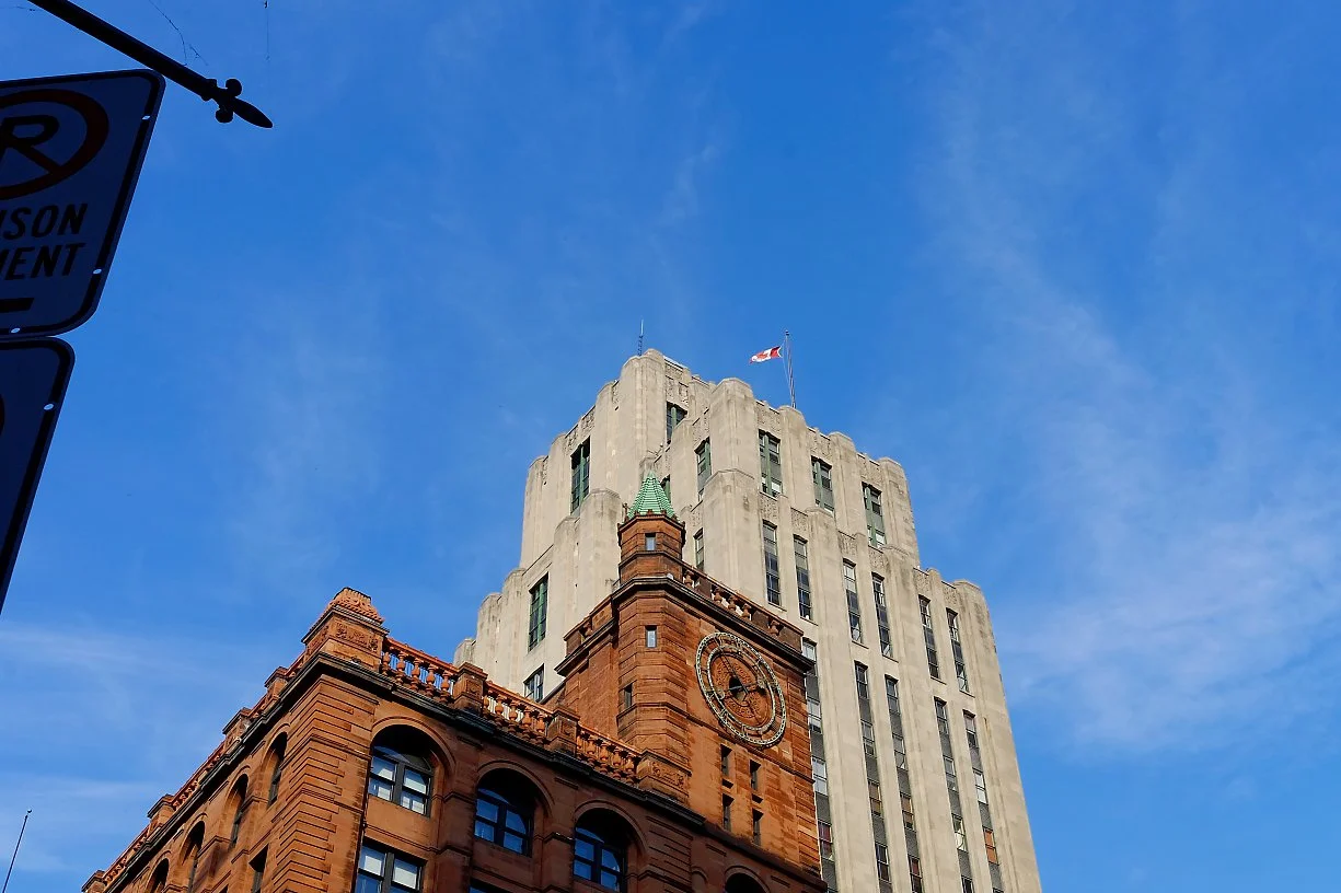The image shows a tall, historic building with a clock tower, with a flag flying on top, set against a bright blue sky. Part of a street sign is visible in the upper left corner.