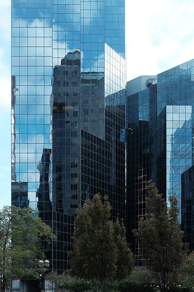 Reflections of nearby buildings on the glass facade of a modern skyscraper, with trees in the foreground.