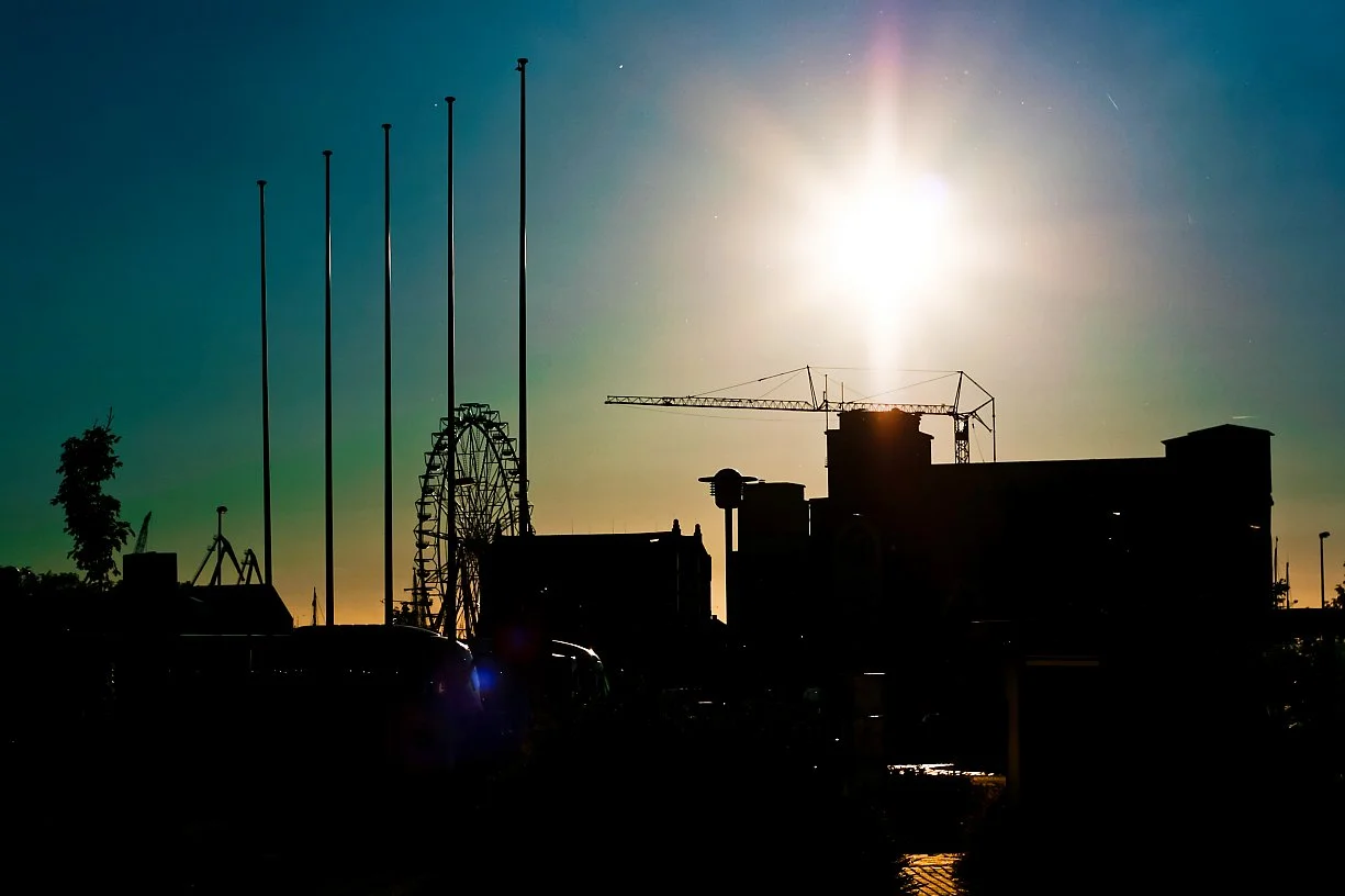 Silhouettes of buildings, a Ferris wheel, and a crane against a bright sun in the sky.