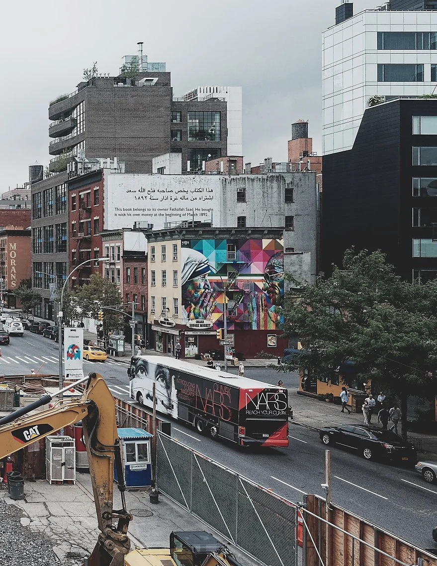 City street scene with buildings, a billboard, a colorful mural, a large bus, and pedestrians walking on the sidewalk.