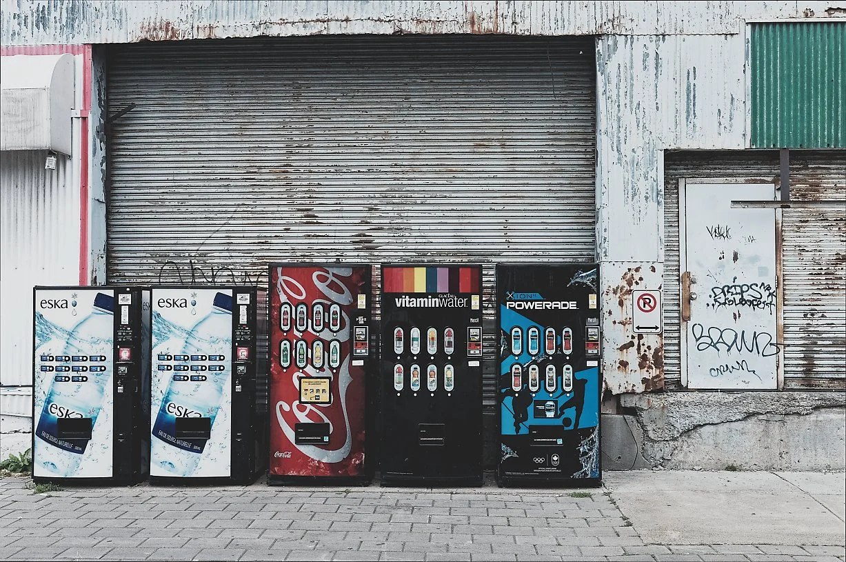 Four vending machines selling drinks are placed outside a weathered building with graffiti and a closed metal rolling door.