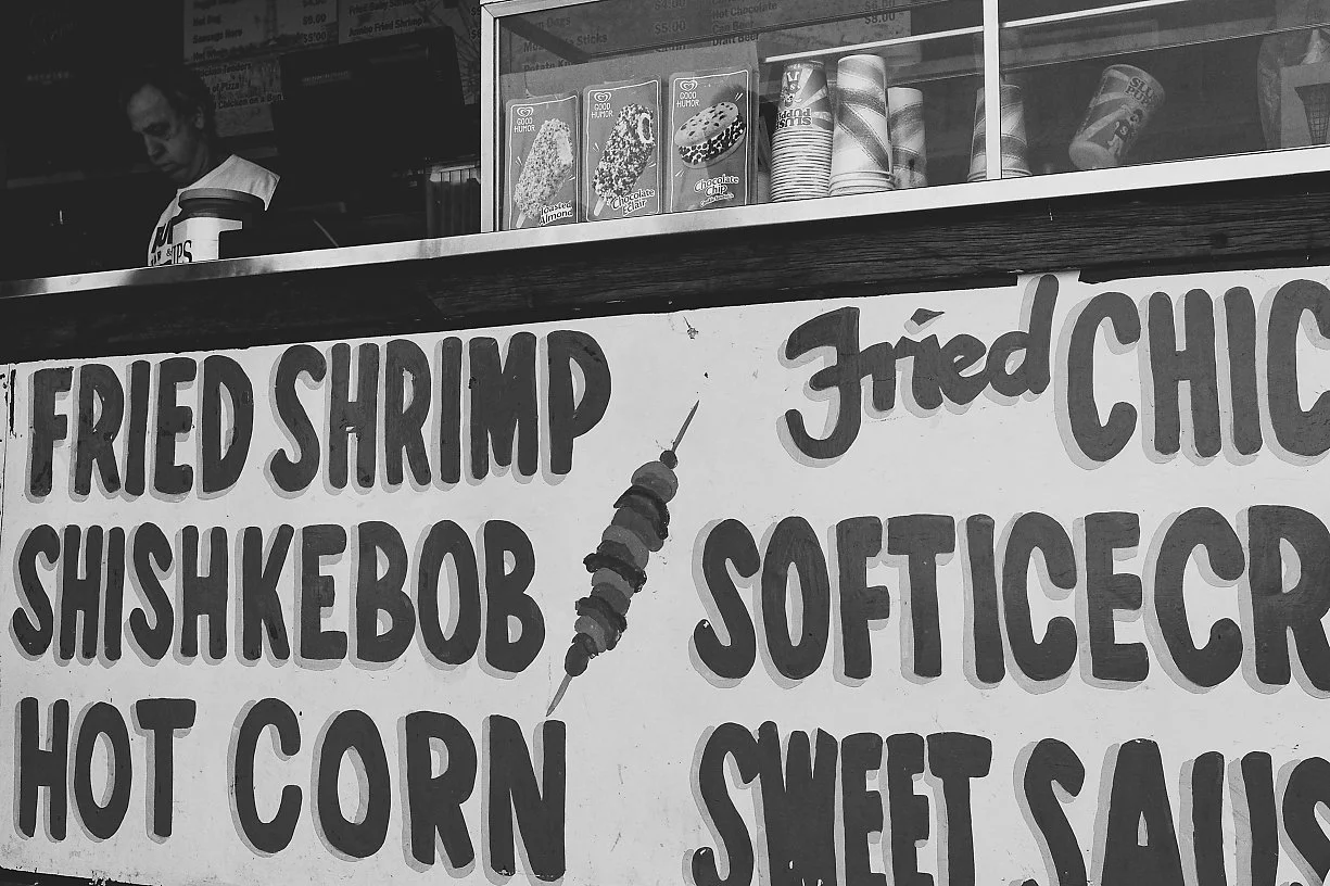 A food stand sign listing fried shrimp, fried chicken, shish kebab, soft ice cream, hot corn, and sweet sausage with a skewer graphic in the center.