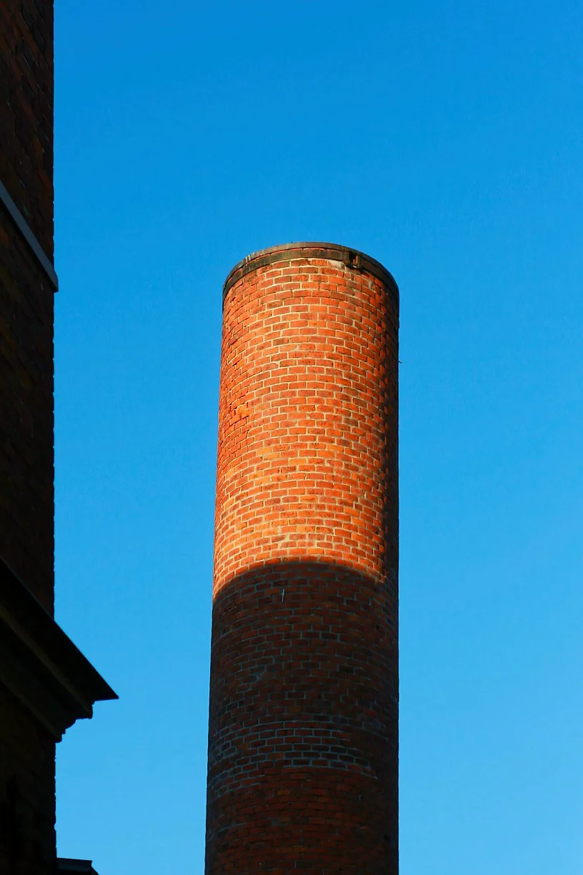 Close-up of a tall, cylindrical brick chimney against a clear blue sky, with part of a building visible on the left.