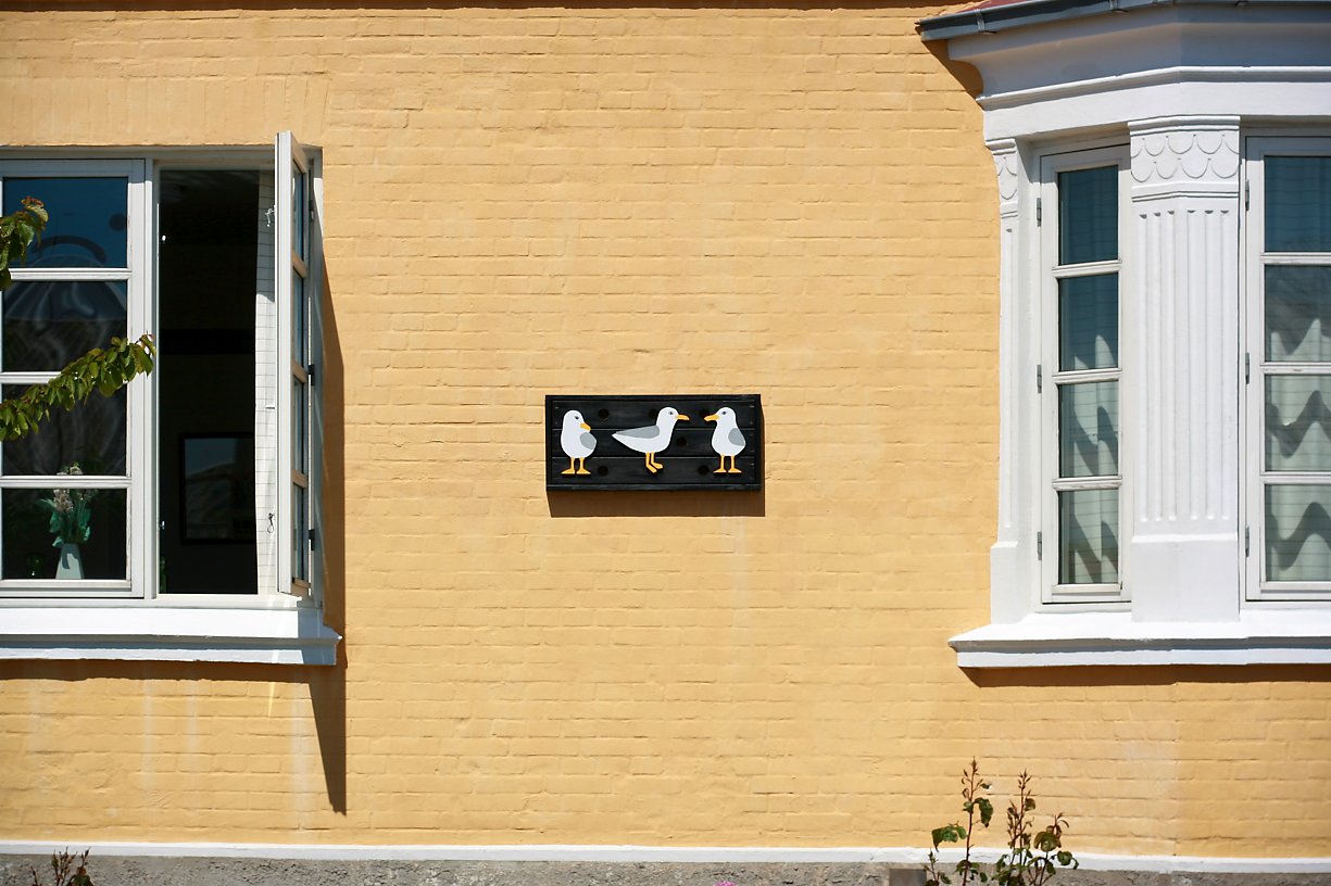 A yellow brick house wall with two windows, one on the left with open shutters and one on the right with closed shutters. In the center of the wall, there's a decorative sign with three cartoon seagulls on a black background.
