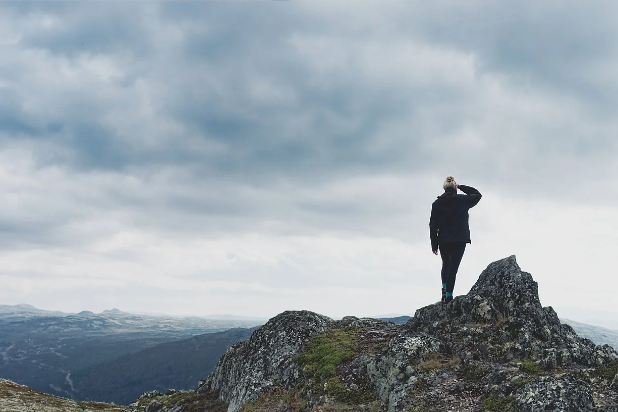 En person står på en fjellknaus og ser ut mot horisonten under en overskyet himmel.