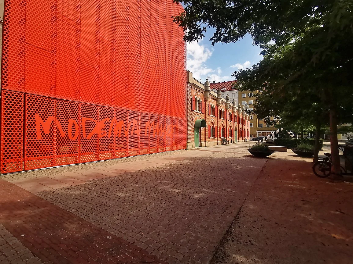 En gate med en stor rød murvegg med perforeringsmønster og graffiti, og en rekke historiske bygninger med buede vinduer ved siden av, omgitt av trær og benker.