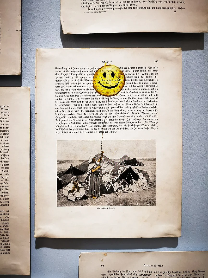 Dette er et svart-hvitt fotografi av en kvinne som holder en ballong med en smiley ansikt malt på den, som flyter over en leir med telt. Flere personer, inkludert barn, er i leiren, og en av kvinnene er sittende på bakken.