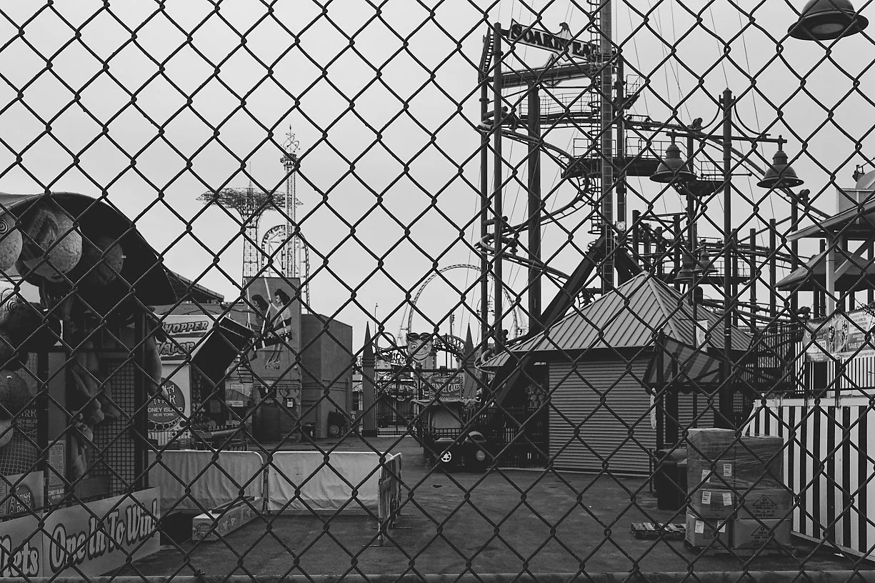 Black and white photo of a deserted amusement park or fairground, viewed through a chain-link fence, with rides, stalls, and signs in the background.