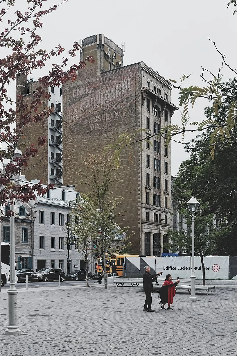A large brick building with faded advertising painted on the side, surrounded by trees and city street with cars and people, on a cloudy day.