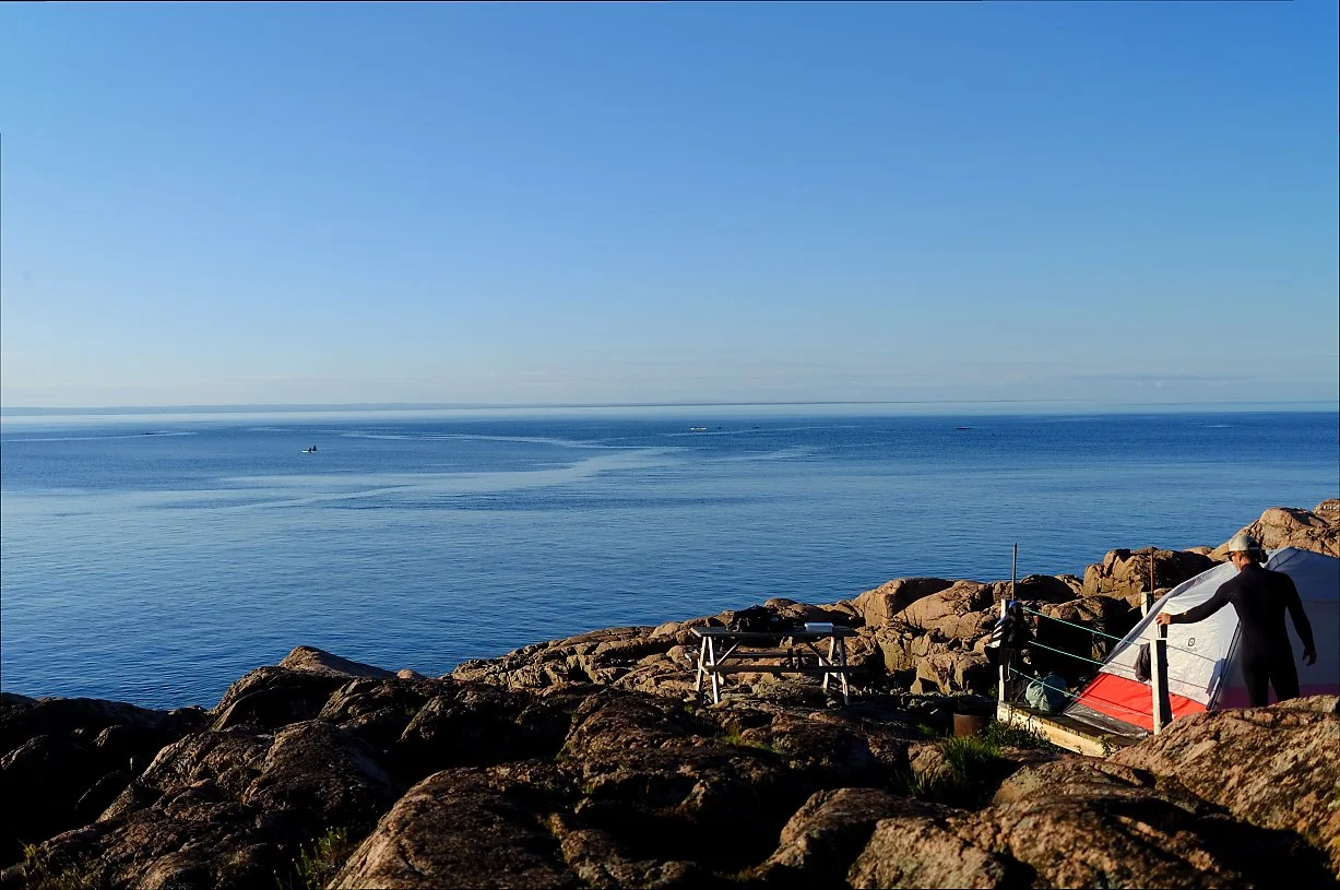 Utsikt over hav med steinete kystlinje og en person som setter opp en telt like ved vannet.