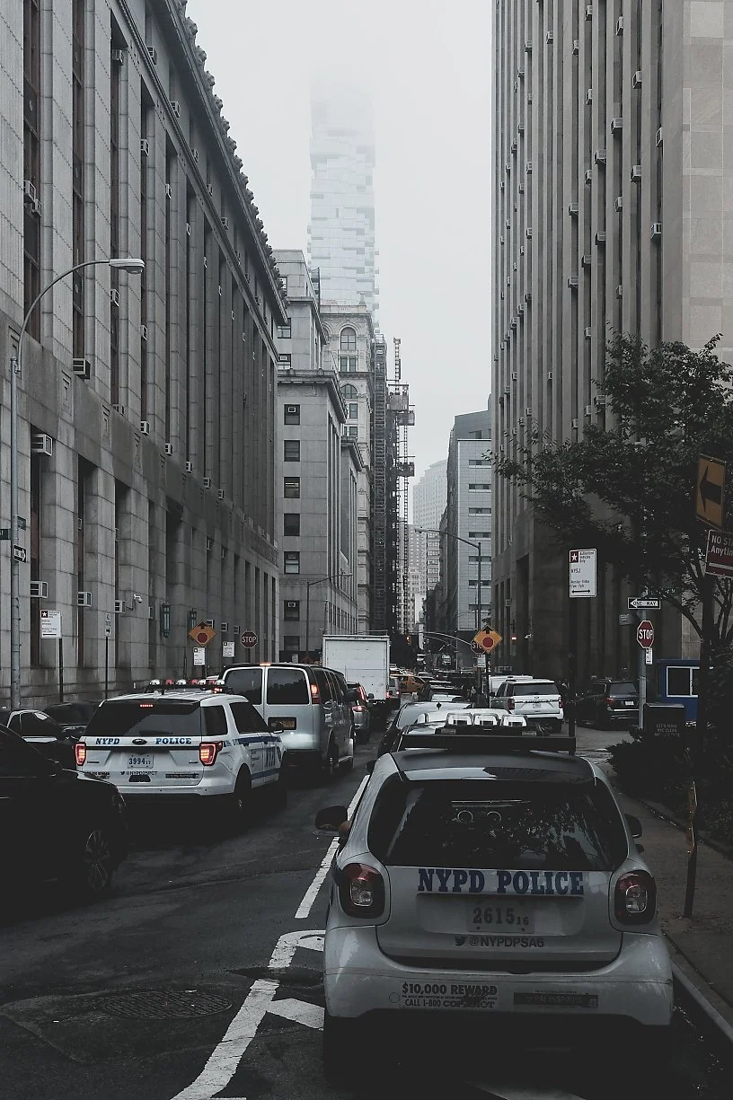 A city street scene with multiple parked and moving vehicles, including two NYPD police cars, surrounded by tall buildings in a gray, overcast weather.