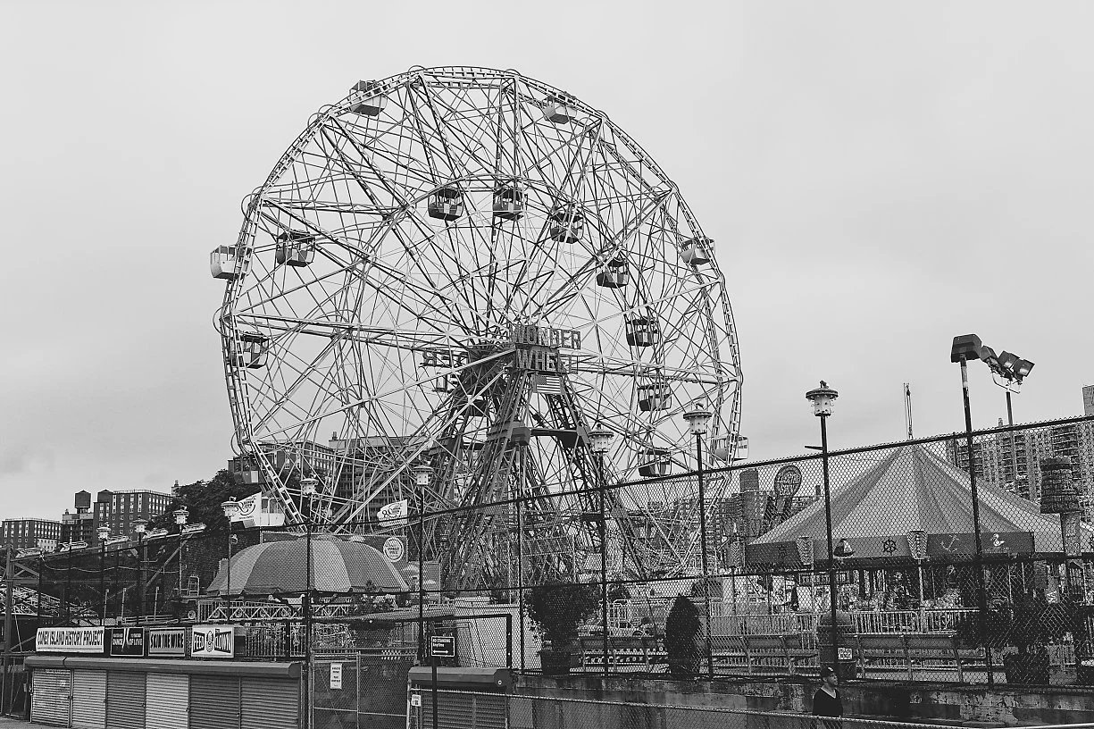 A black and white photo of a large Ferris wheel at an amusement park with a fenced area in the foreground and a city skyline in the background.