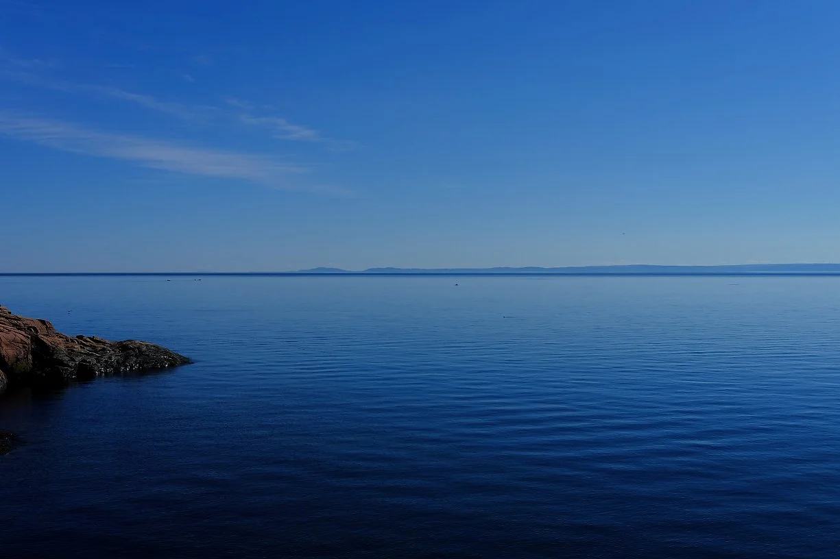 Et rolig hav med blå vann og en klar, overskyet himmel. En steinstrand i venstre hjørne og en fjern horisont med lave fjell eller øyer.