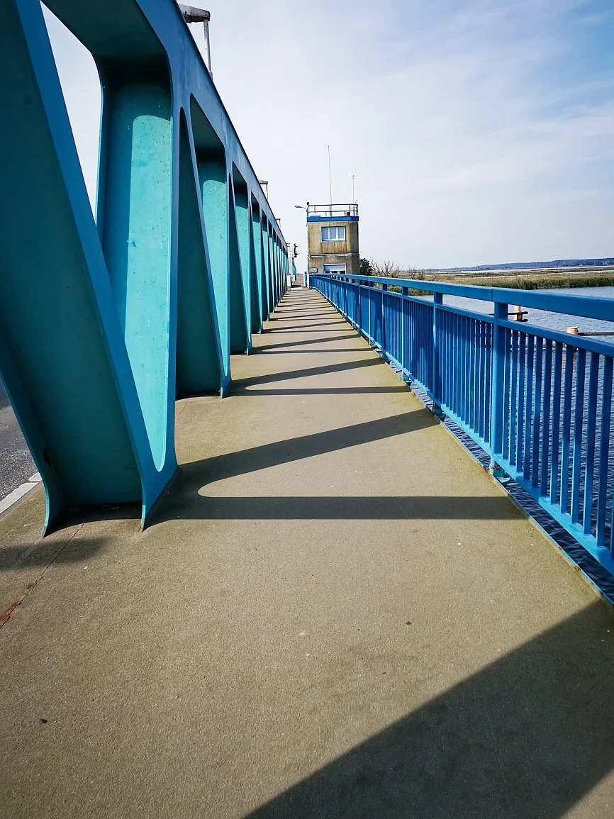 A walkway with a blue railing alongside a body of water, with a small control tower or observation building at the end of the walkway. The sky is clear with some light clouds.