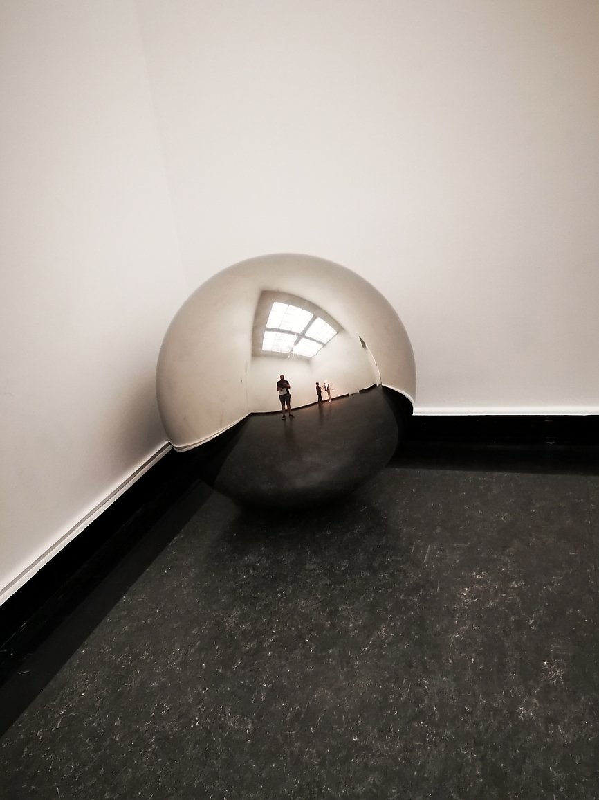 Refleksjon av to personer i en stor, rund, speilende skulptur plassert på gulvet i et rom med hvite vegger og store vinduer.