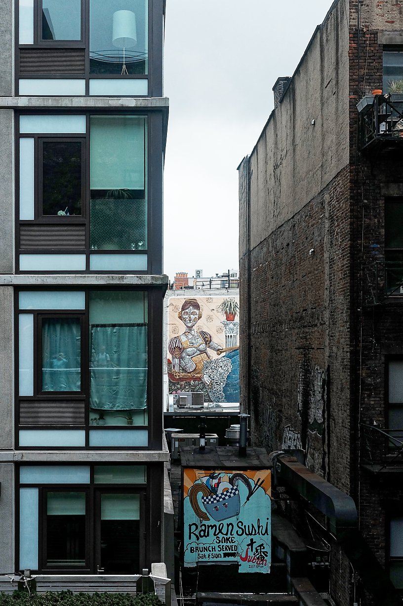 View of tall, modern apartment building on the left, with large windows and curtains inside, on a cloudy day. To the right, an older brick building with fire escape. In the background, a colorful mural of a woman playing guitar is visible on a distant building wall. A sign for a ramen and sushi restaurant called Sakura, offering brunch on Saturdays and Sundays, is at the bottom center of the image.