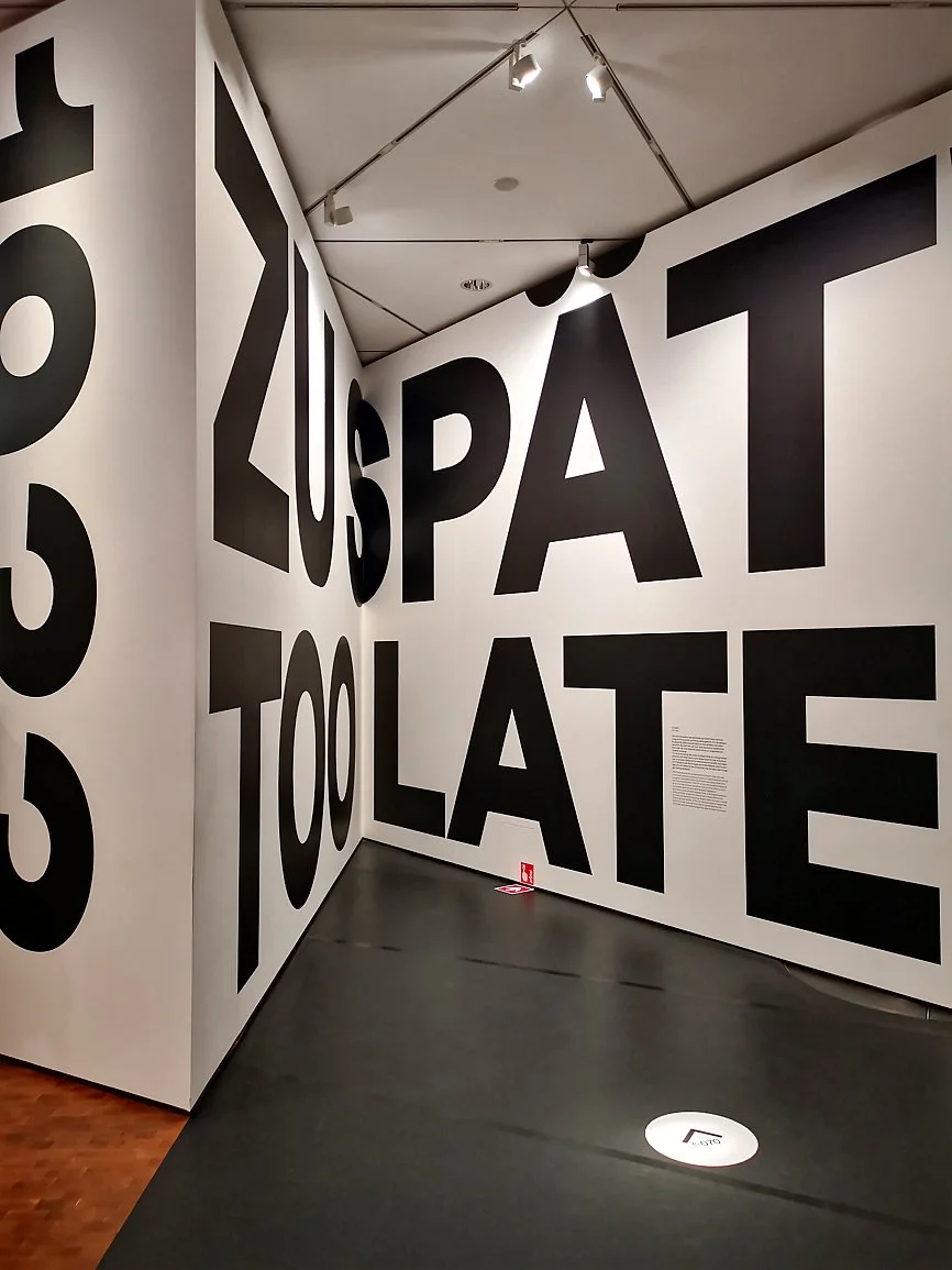 Interior of an art exhibit with large black letters on white walls spelling out words like "US PAT", "TOOLATE" and "2023" in a modern gallery setting with track lighting.