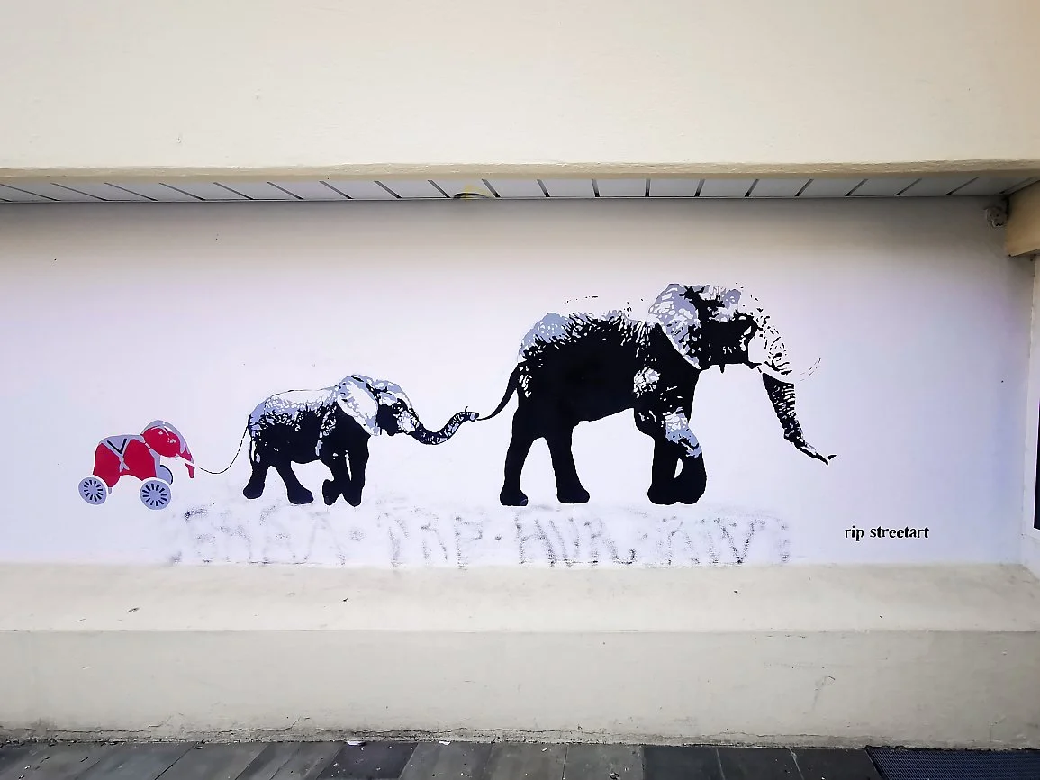 Veggmaleri av tre elefanter, de minste drar de større, på en hvit vegg. Det er også en liten sykkelfigur på venstre side av bildet.
