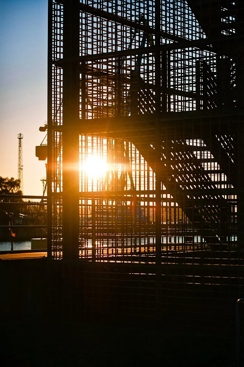 Sunset behind a large metal scaffolding structure with stairs and grid pattern, with a body of water and trees in the background.
