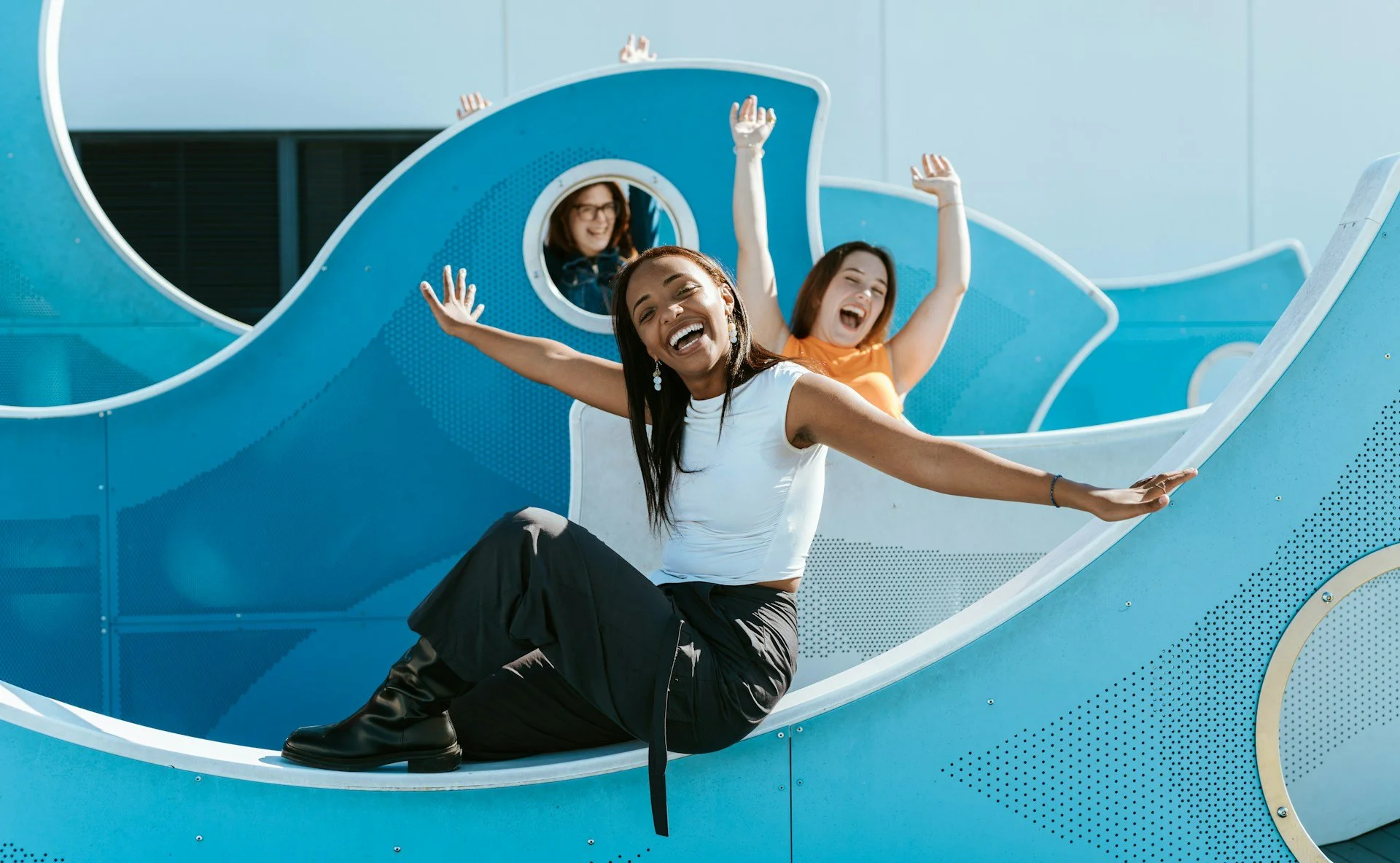 Three female teens are riding a blue, modern, and artistic slide at a playground, laughing and enjoying themselves.