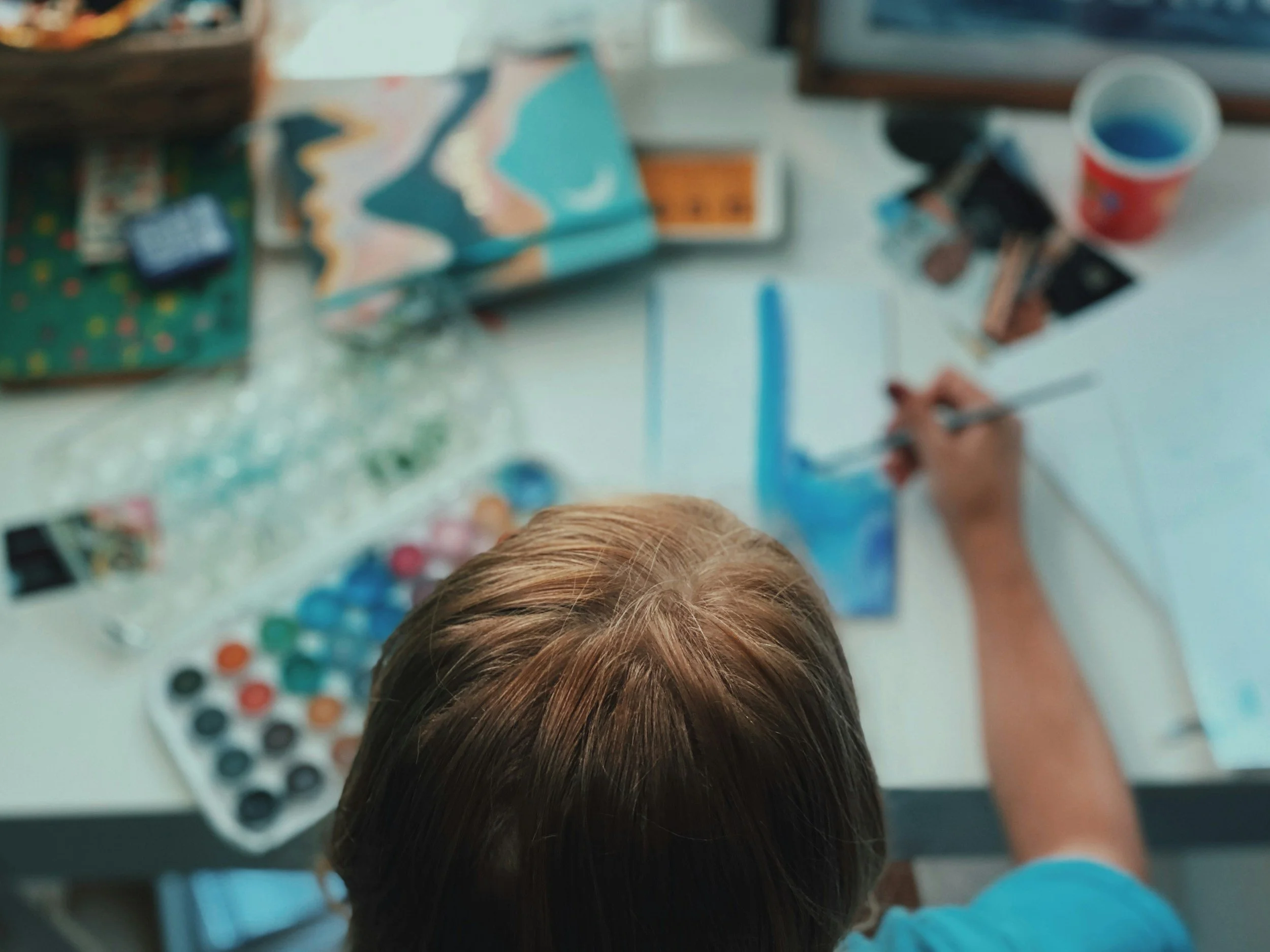 Child painting a picture with blue watercolor paint at a white table surrounded by art supplies and a cup.