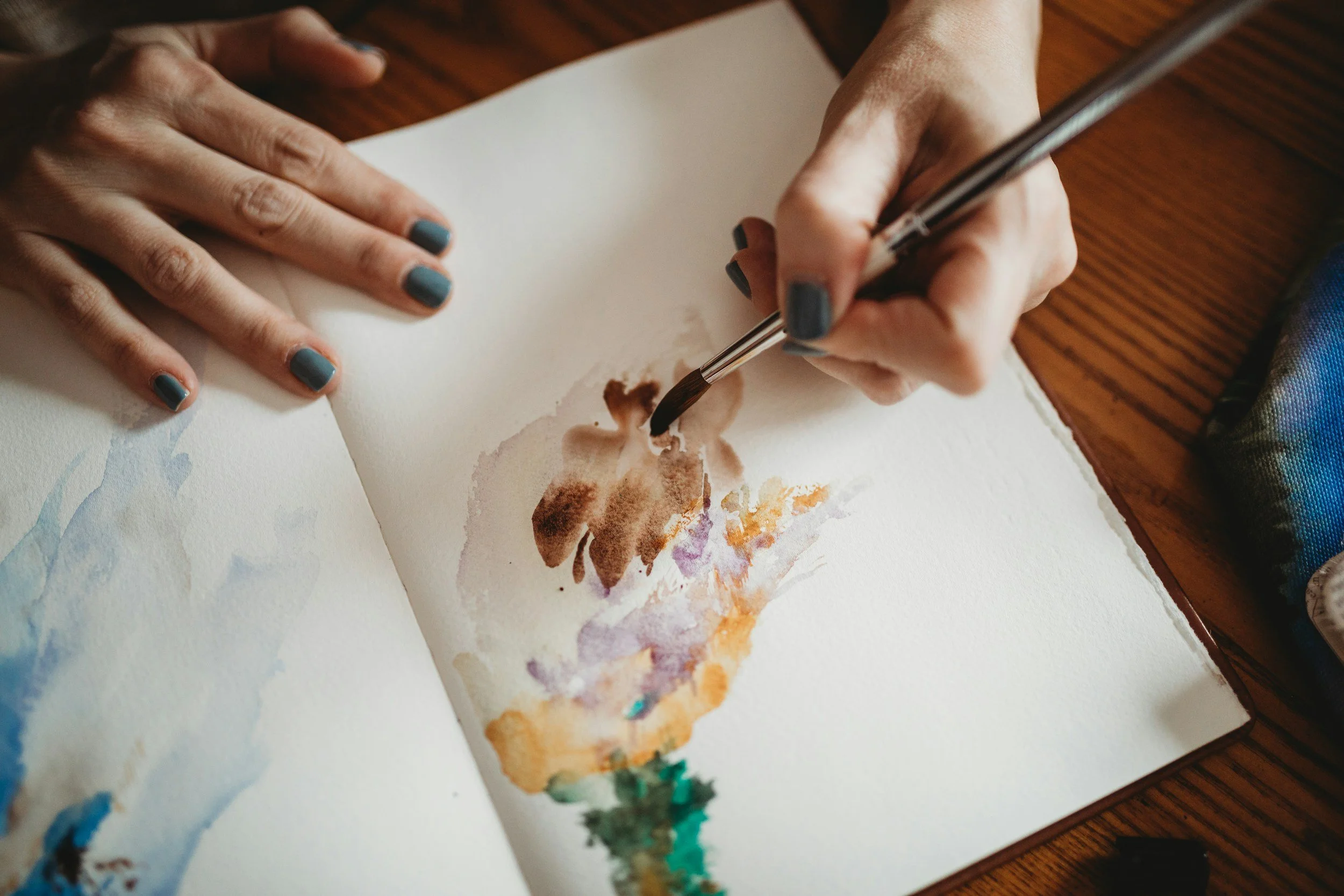 Person painting with watercolors on a sketchbook, using a brush, with hands visible and a wooden table surface.