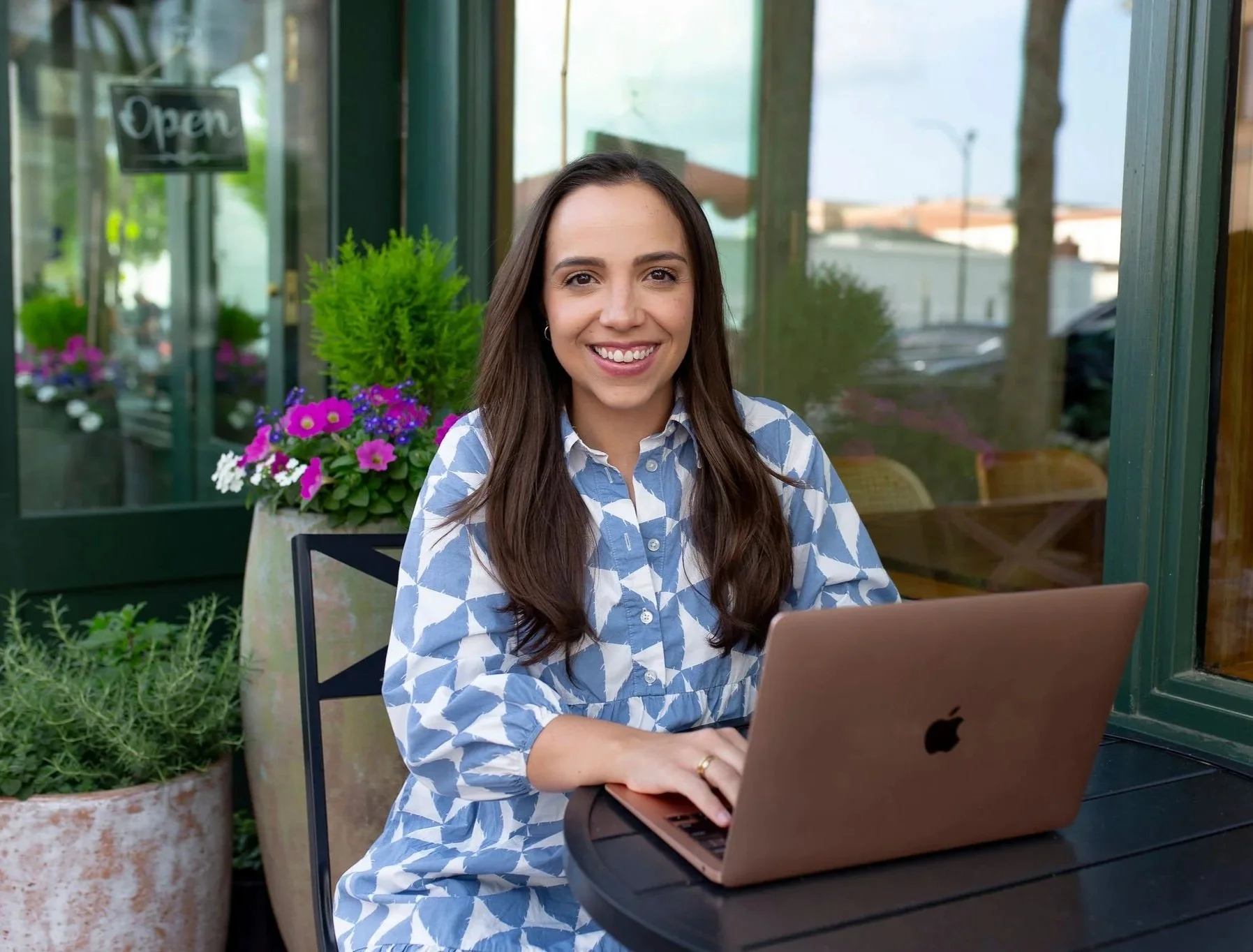 Woman sitting at an outdoor table working on a laptop, smiling, with colorful flowers and potted plants in the background.