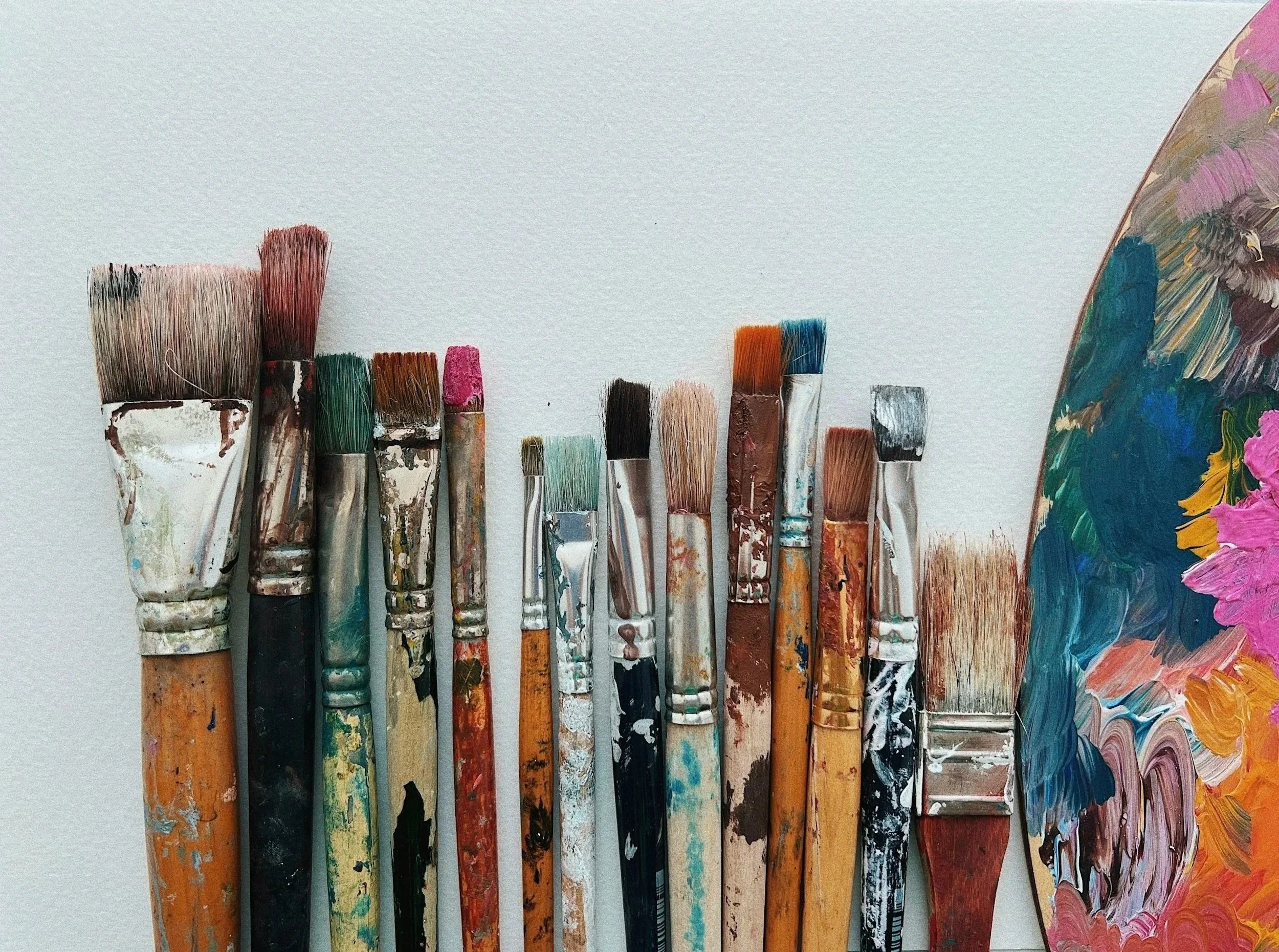 Collection of used paintbrushes with paint splatters and a colorful palette on the right side, all positioned vertically against a plain light background.