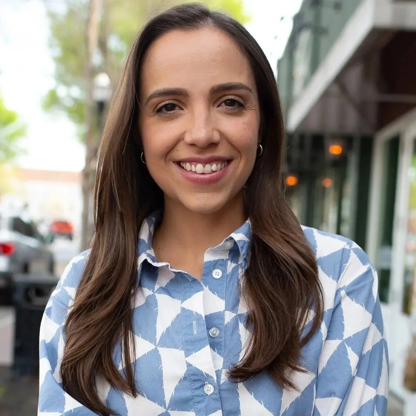 A young woman with long dark brown hair smiling outdoors, wearing a blue and white patterned button-up shirt.
