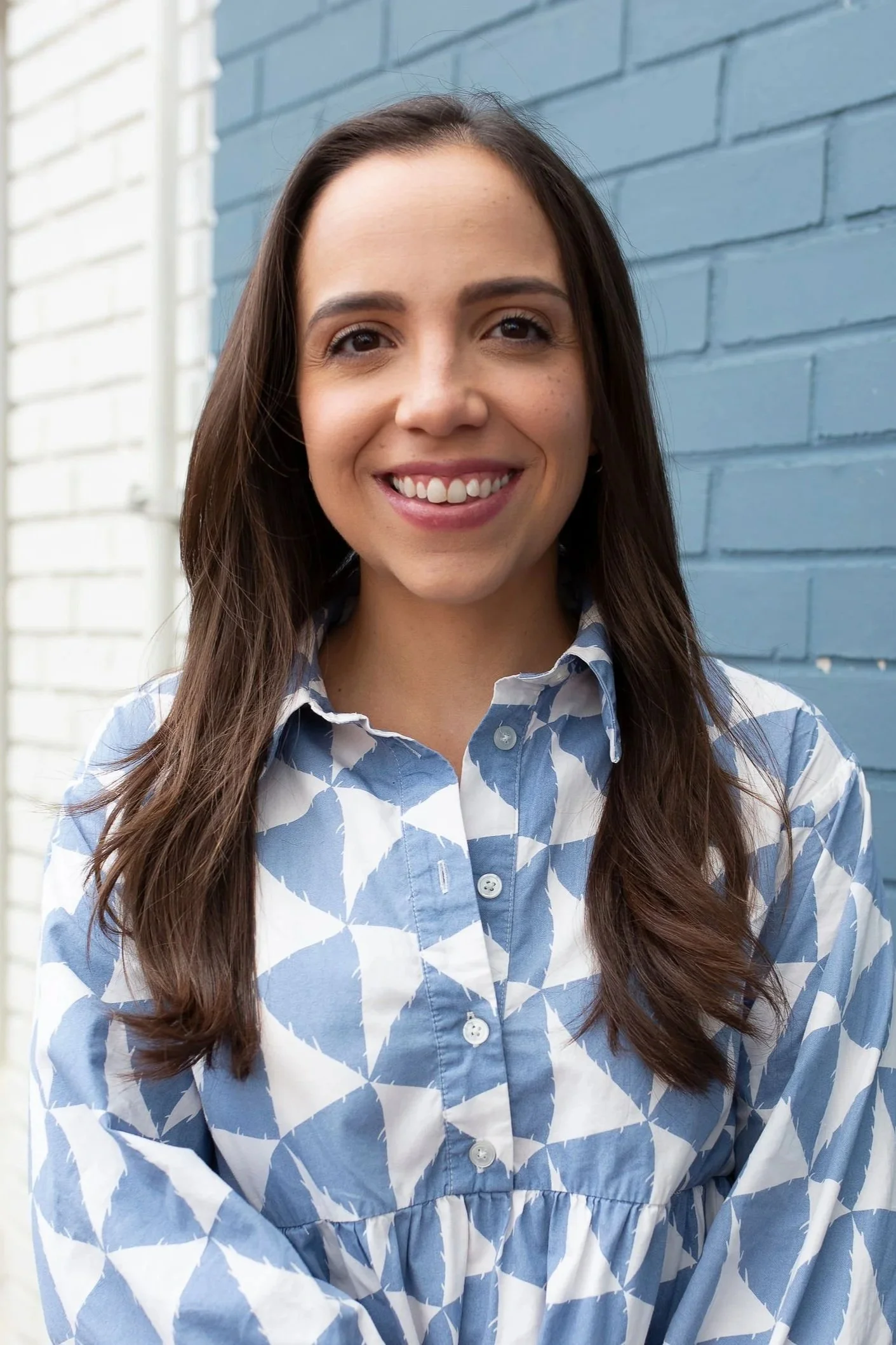 A woman with long brown hair, smiling, wearing a blue and white patterned button-up shirt, standing in front of a blue brick wall.