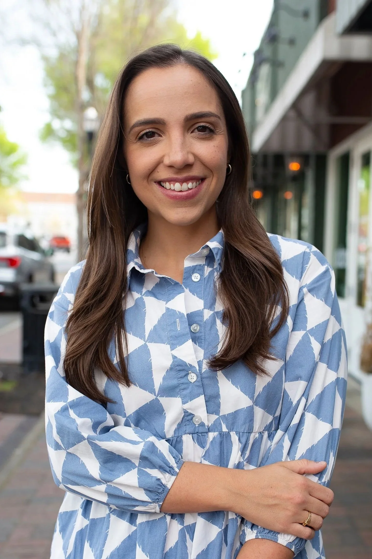 A young woman with long brown hair, smiling, wearing a blue and white patterned button-up dress, standing outdoors in front of a storefront with trees and cars in the background.