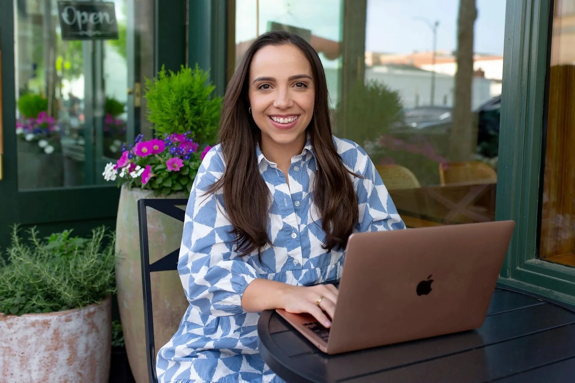 woman in a blue and white dress sitting at a table smiling and typing on laptop